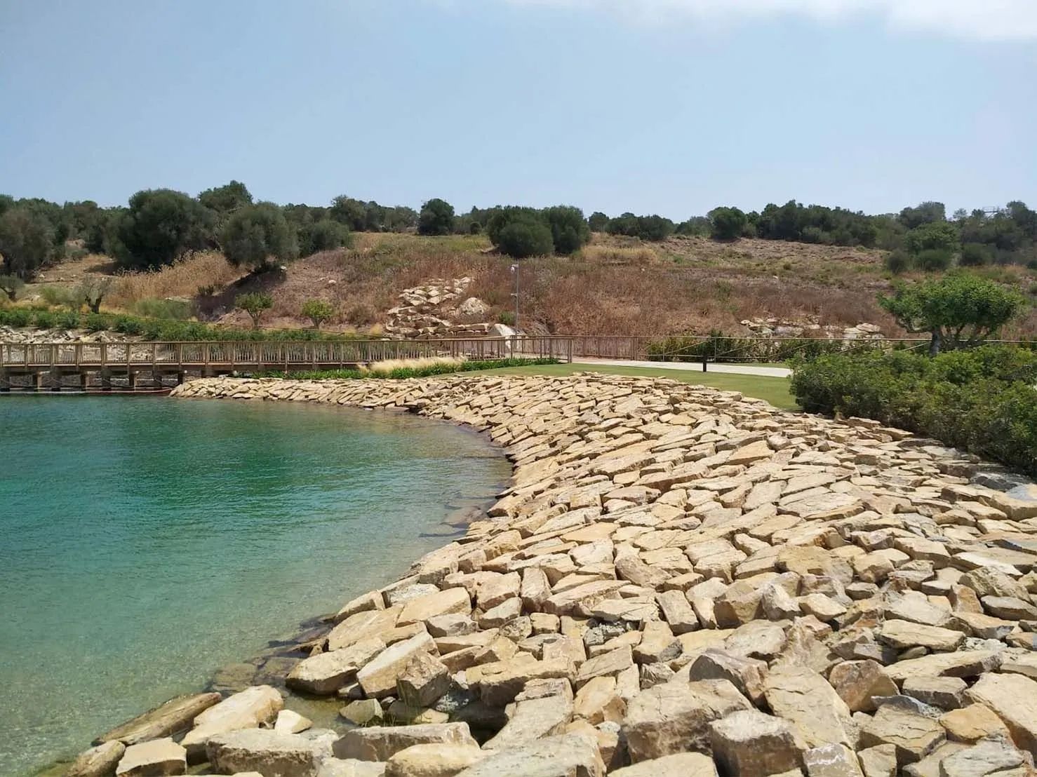 Costa bordeada de piedras, con aguas turquesas, un puente y una colina cubierta de hierba bajo un cielo azul.