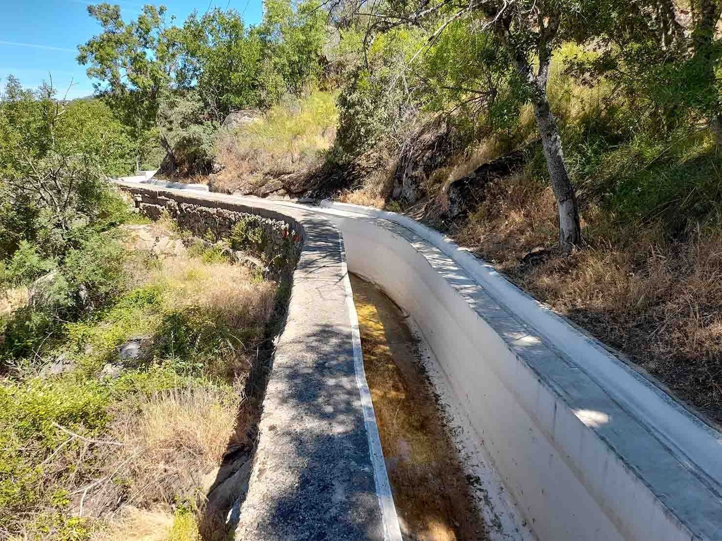 Canal de riego de hormigón que serpentea por la ladera de una colina, rodeado de árboles y vegetación seca.