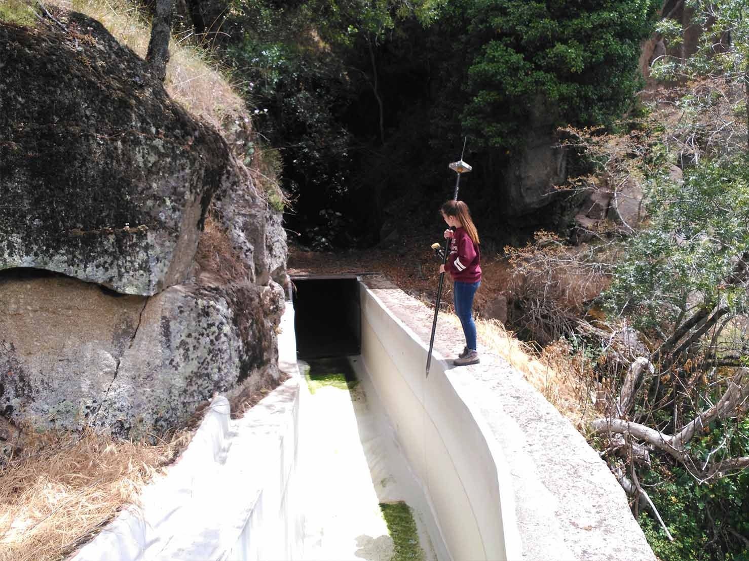 Mujer de pie sobre un canal de hormigón, usando un dispositivo GPS cerca de la entrada de un túnel oscuro. Acantilado rocoso a la izquierda, árboles detrás.