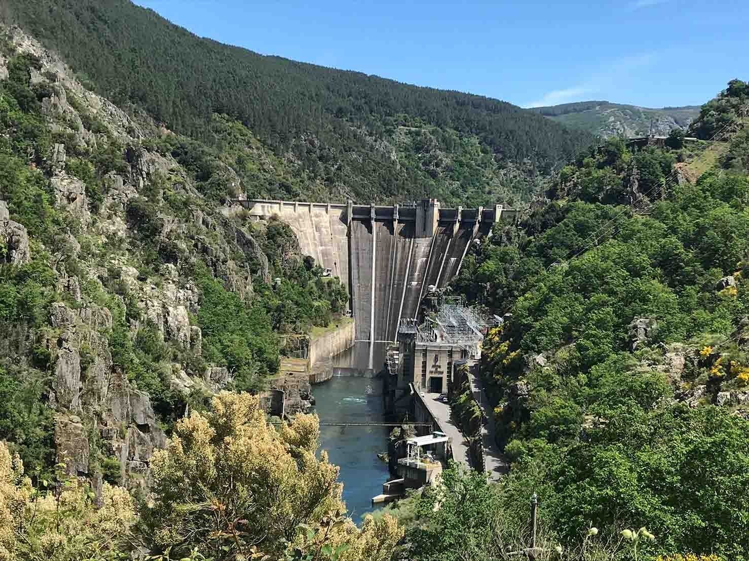Presa en un valle verde, con agua fluyendo sobre ella bajo un cielo azul.