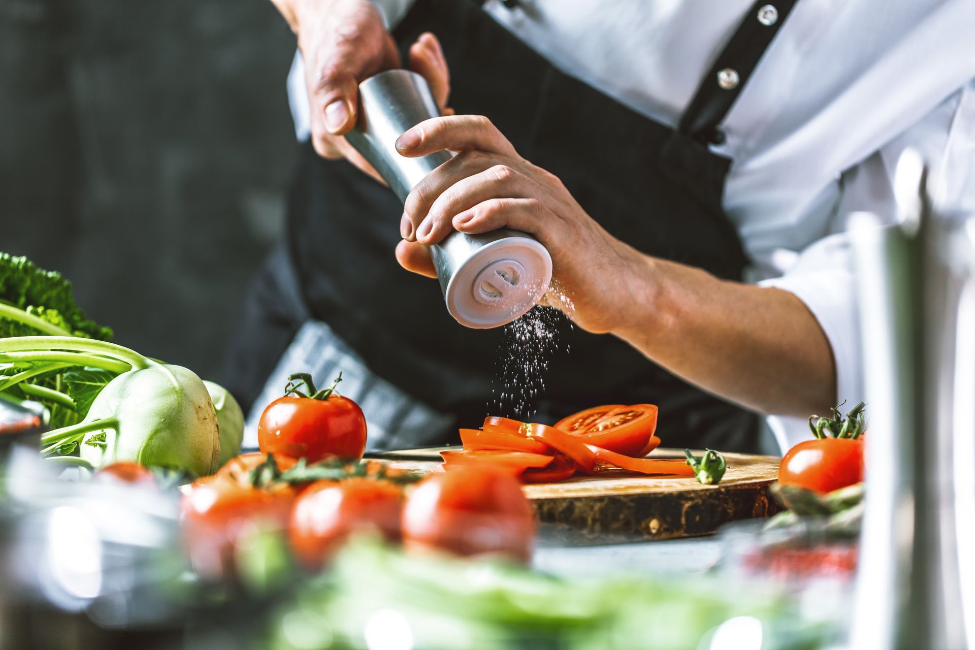 Dans une cuisine, un chef utilise un moulin à épices pour assaisonner des tomates coupées sur une planche à découper en bois.