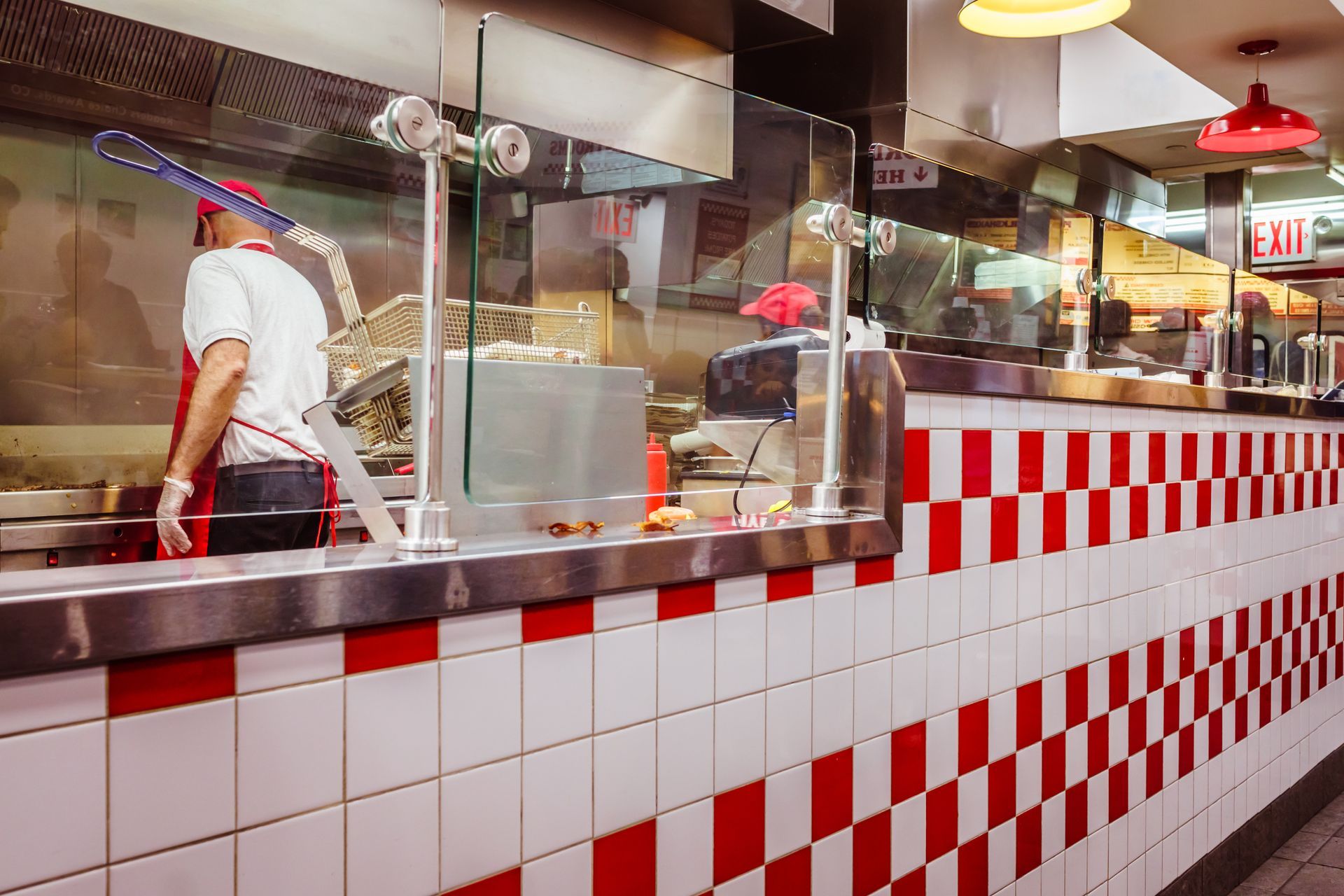 Dans un restaurant, un employé en uniforme prépare des plats derrière un comptoir décoré de carreaux à damier rouge et blanc.