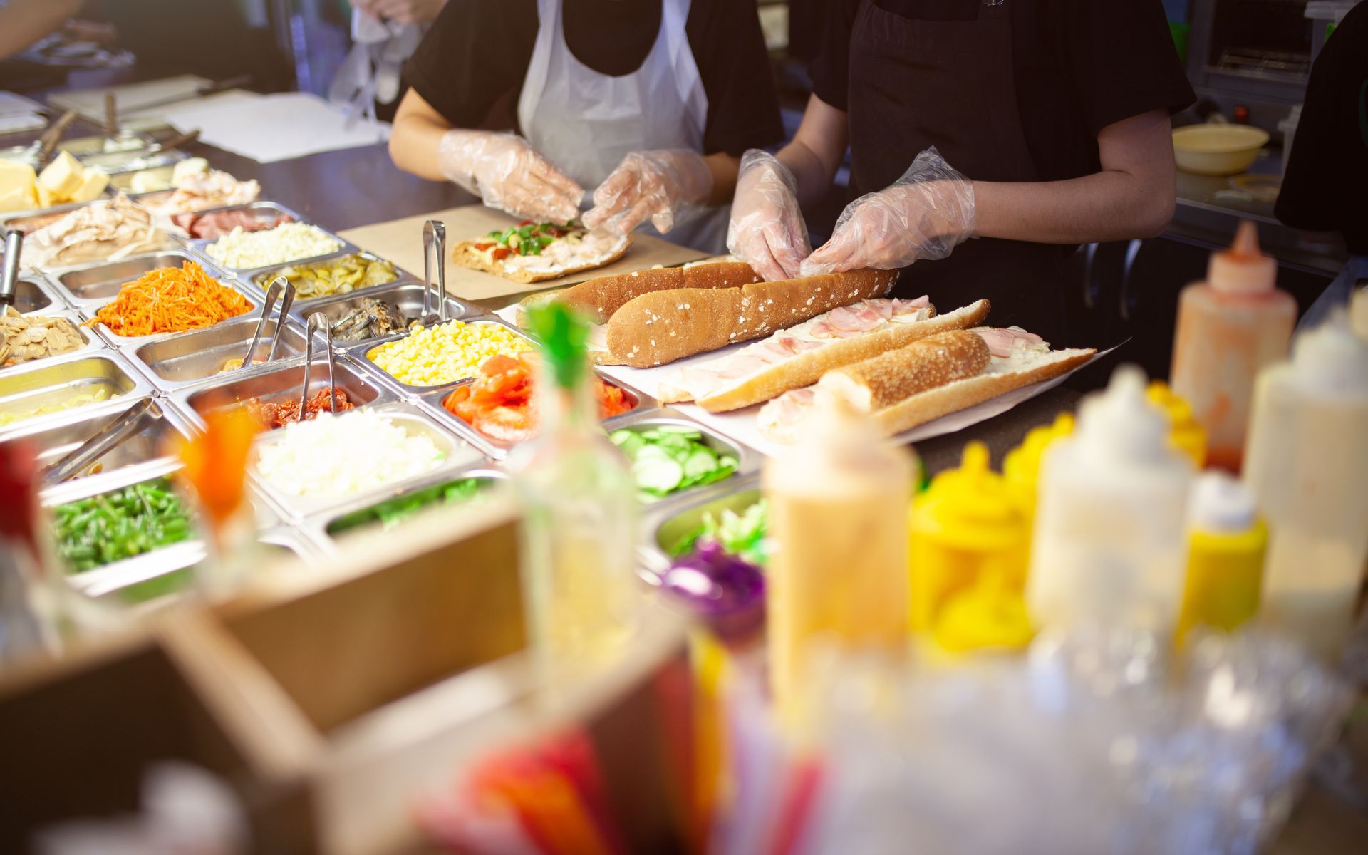 Deux personnes portant des gants préparent des sandwichs à un comptoir de restauration rempli d'ingrédients.