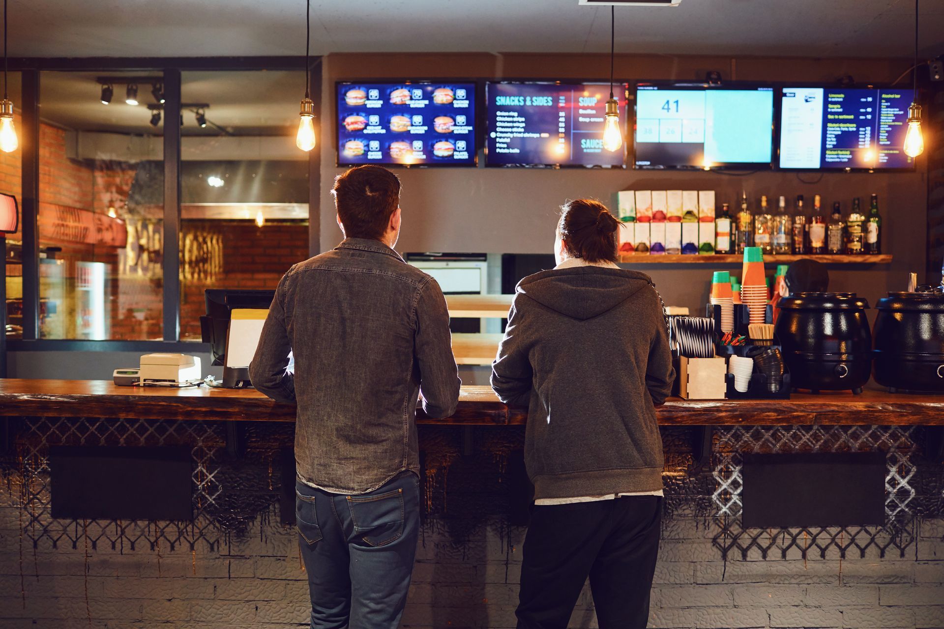 Deux personnes se tiennent debout derrière un comptoir dans un restaurant, face à des menus numériques affichés au mur.