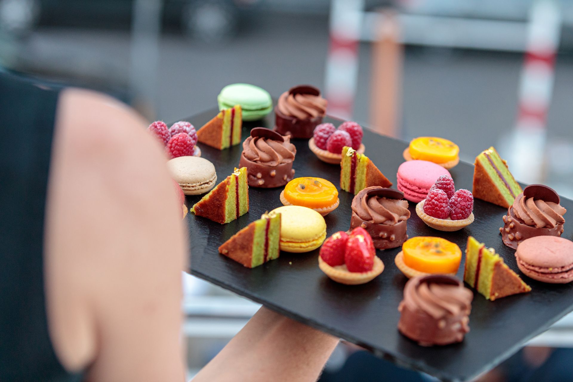 Une personne tient un plateau rempli d'assortiment de macarons, de tartelettes au chocolat et de pâtisseries aux fruits.