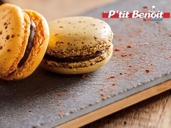Deux macarons beiges fourrés au chocolat noir, posés sur une surface en ardoise saupoudrée de poudre de chocolat.