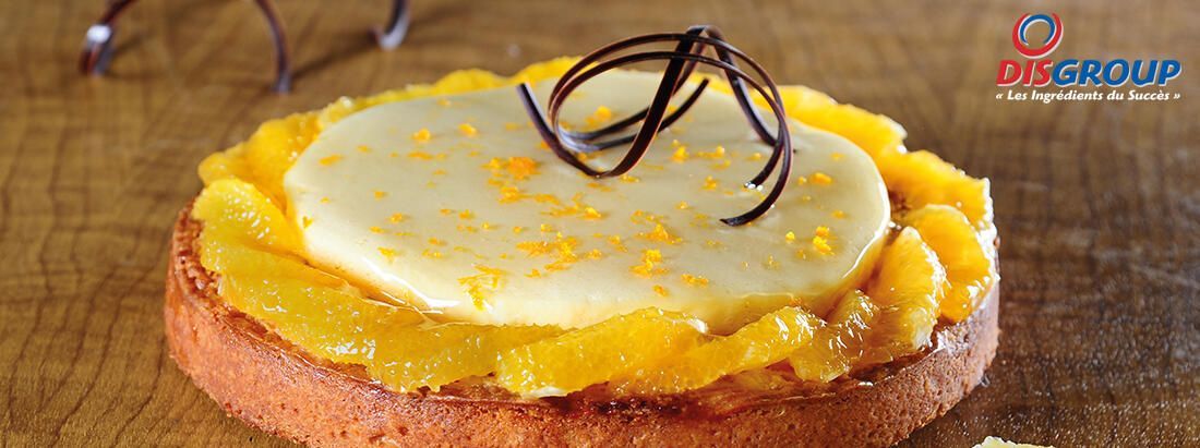 Une tarte à l'orange sur un support en bois, garnie d'un disque de crème, de zestes d'agrumes et de copeaux de chocolat.