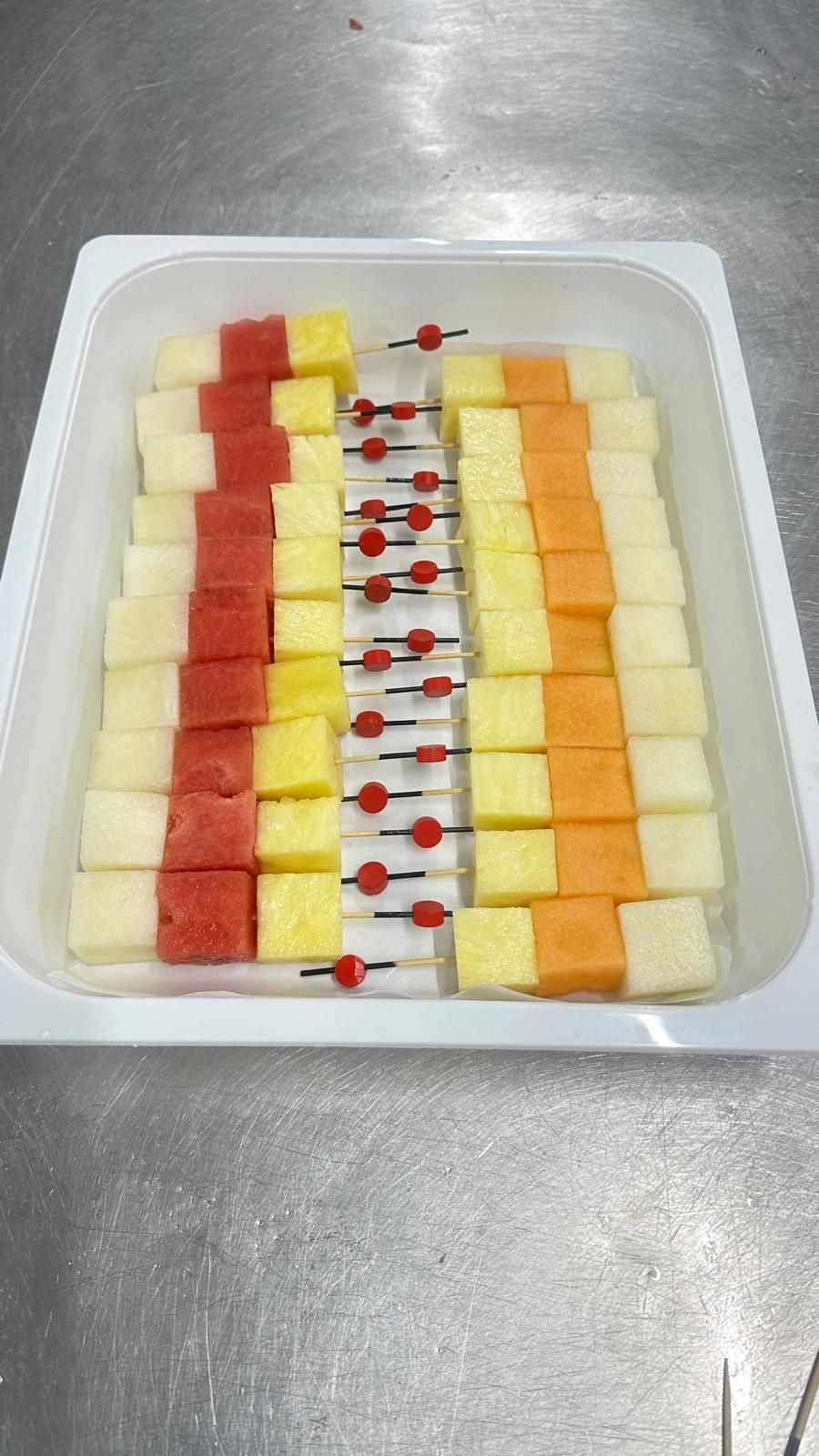 Una bandeja blanca llena de frutas cortadas en brochetas.