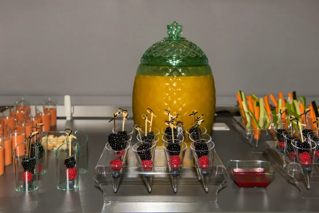 Una mesa de buffet con un dispensador de bebidas en forma de piña y brochetas de frutas.