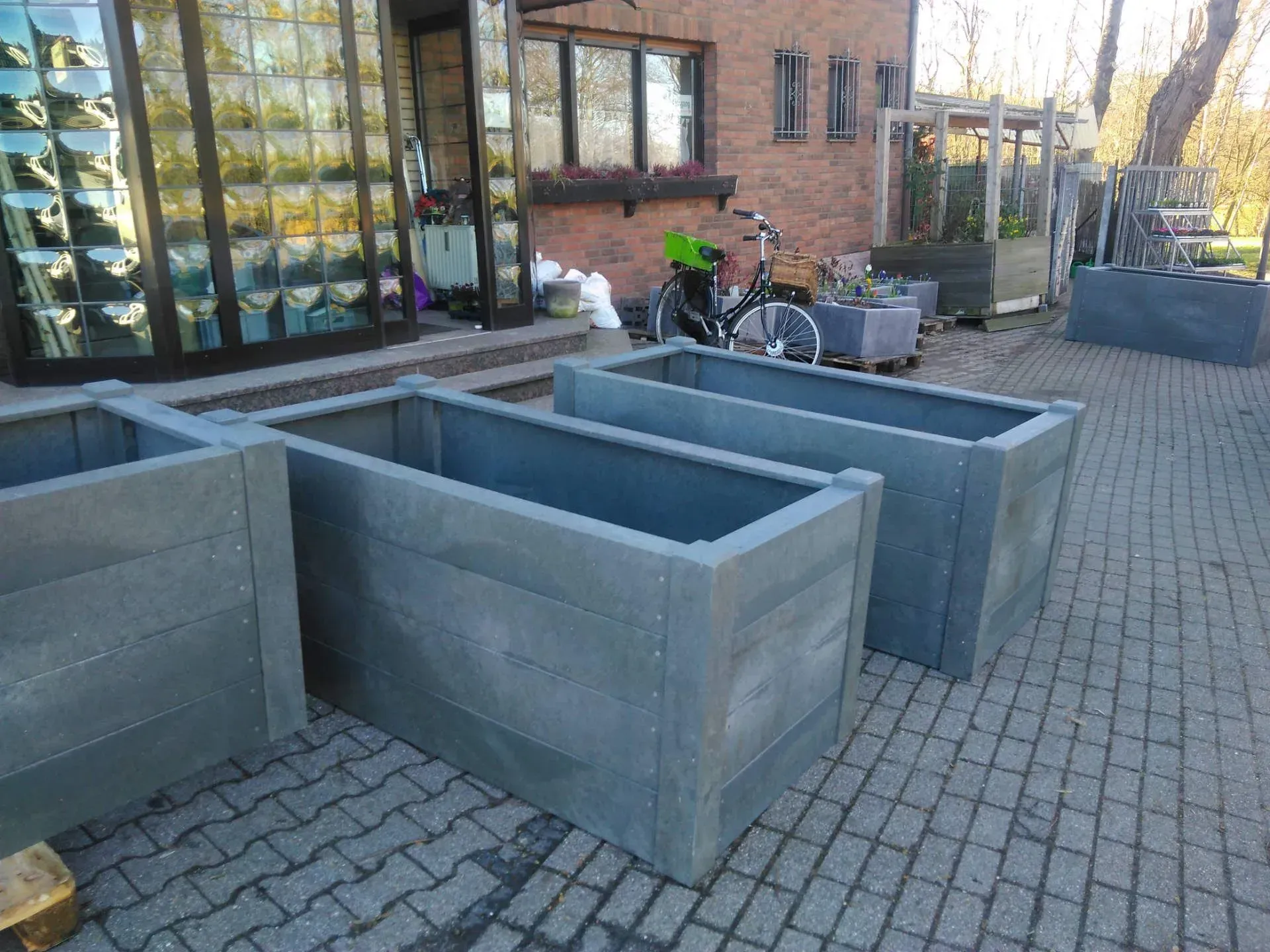 Drei Pflanzkübel im Beton-Look stehen auf einem gepflasterten Gehweg vor einem Haus.