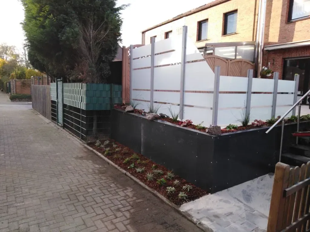 Ein gepflasterter Gehweg, an der Seite ein mit Glaszaun mit Begrünung ringsherum