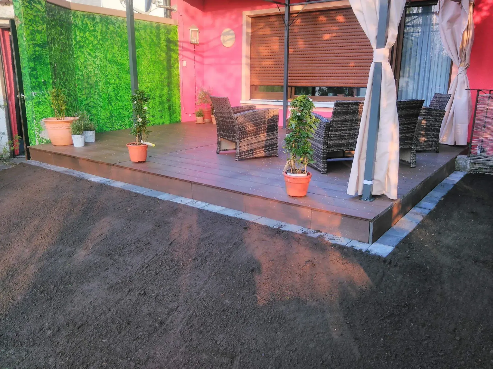 Eine Holzterrasse mit Stühlen und Topfpflanzen vor einem rosa Haus