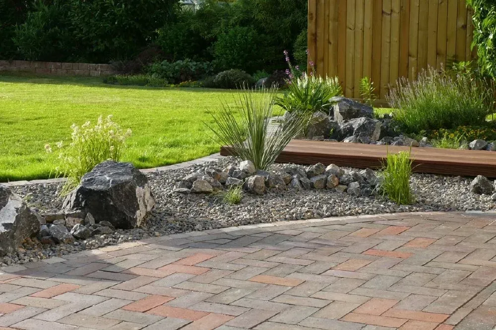 Eine gepflasterte Terrasse, an die ein Steinbeet und einen grünen Rasen grenzt, im Hintergrund ist ein Holzzaun zu sehen.