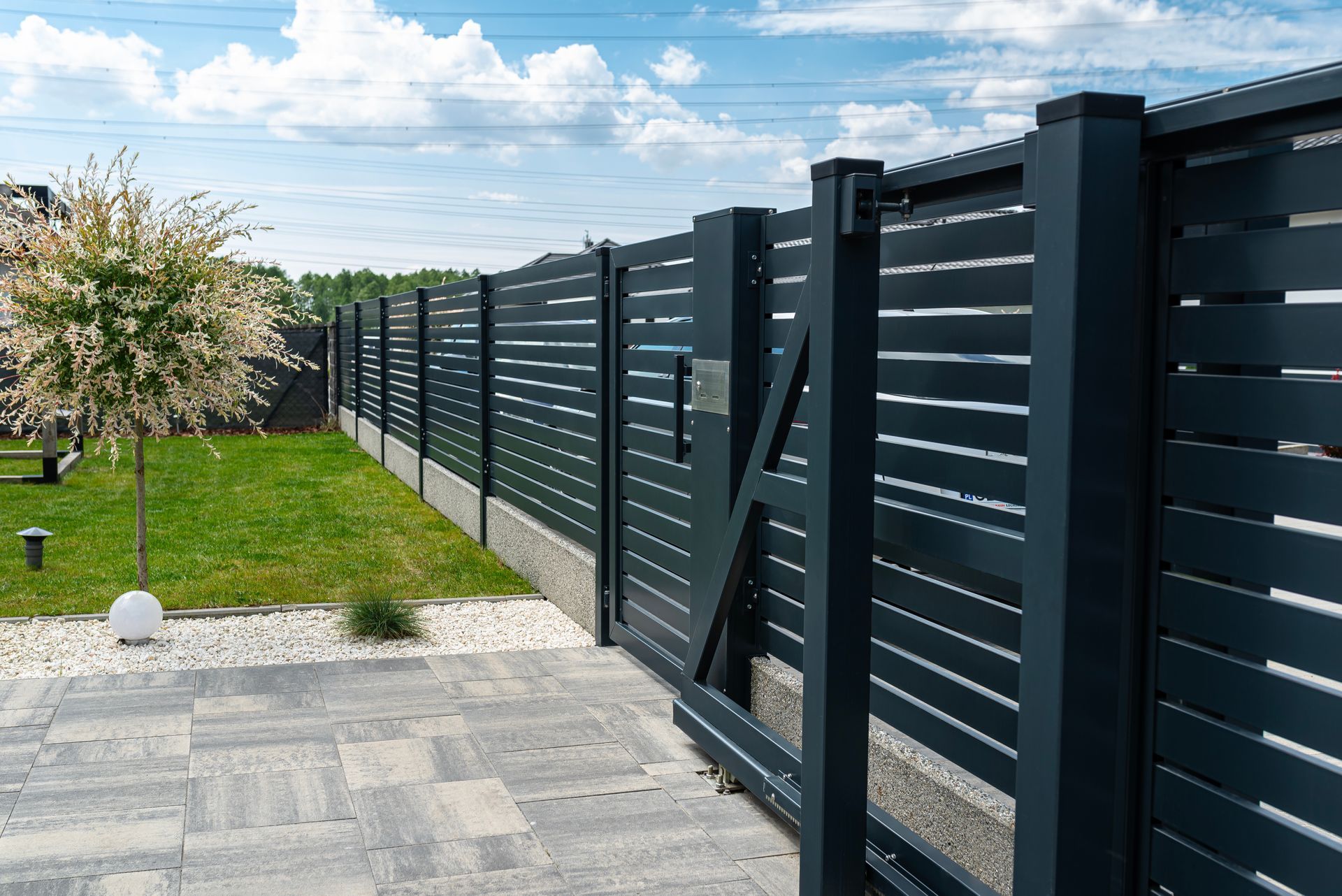 Ein dunkler moderner Zaun mit einem Schiebetor umschließt einen Garten.