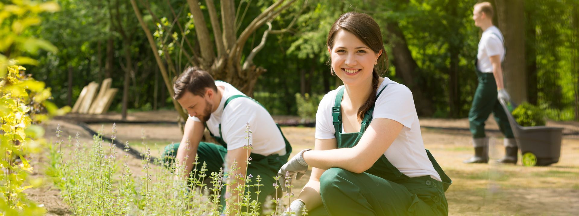 Eine Gärtnerin grinst in die Kamera, hinter ihr hockt ein Gärtner und arbeitet i