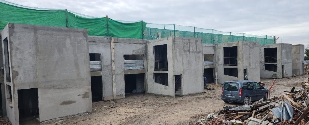 Filas de módulos prefabricados de hormigón sin terminar en una obra. Un coche oscuro está aparcado cerca. Hay una malla verde encima.