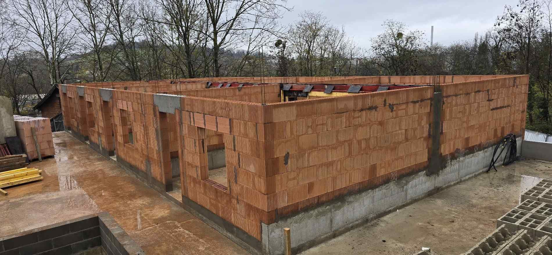 Obra en construcción con paredes de ladrillo visto, ventanas y cimientos de hormigón. Árboles al fondo. Cielo nublado.