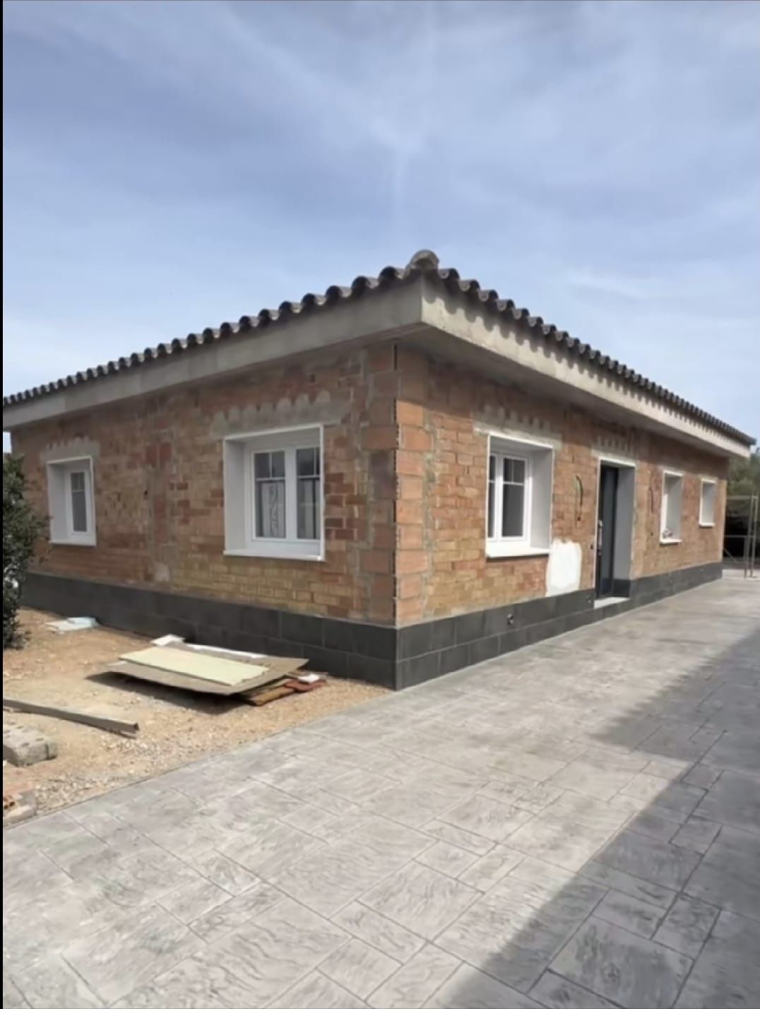 Casa de ladrillo con ventanas de marco blanco, base oscura y techo de tejas sobre un patio de hormigón.