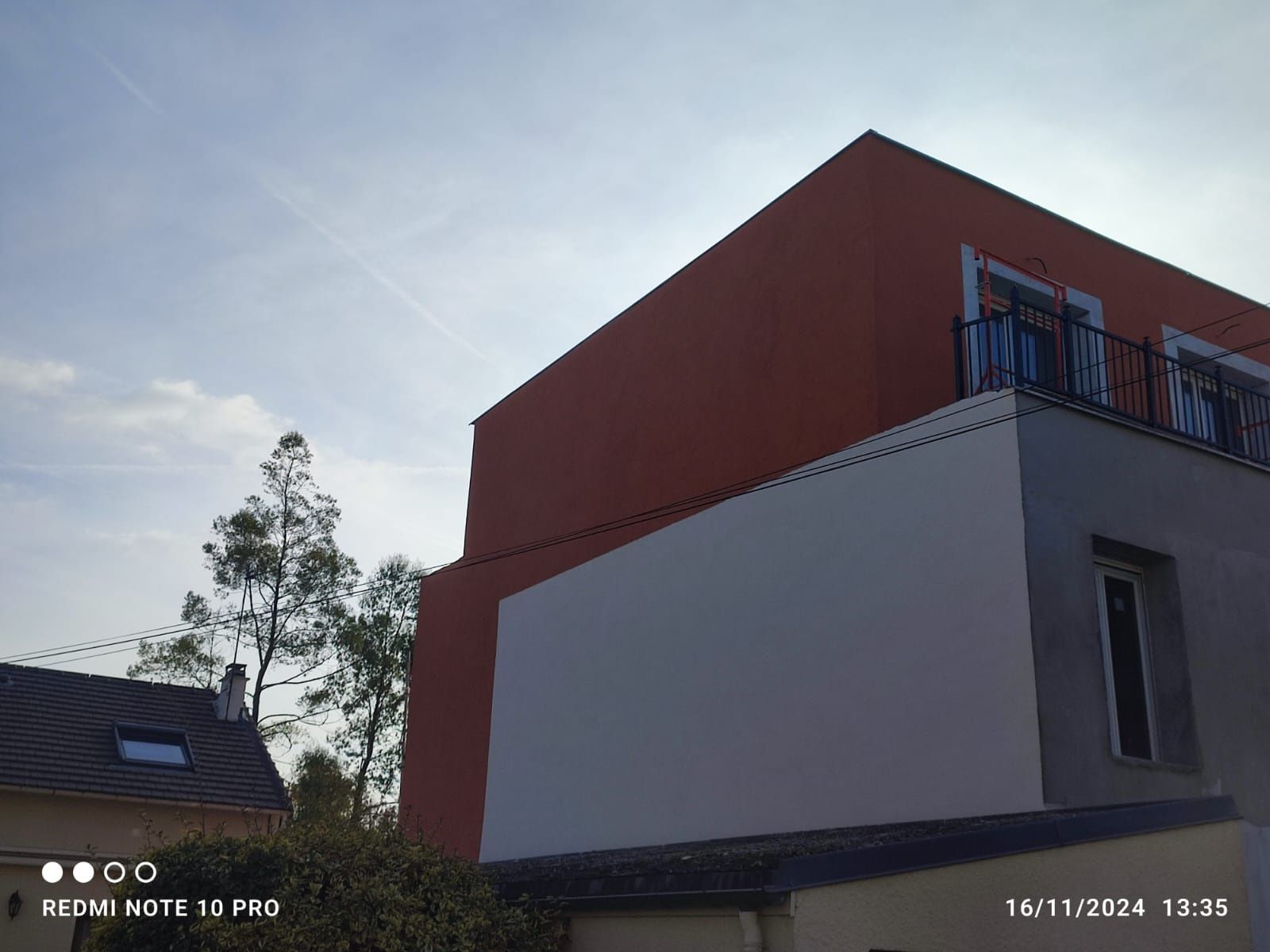 Edificio de dos plantas con paredes rojas y blancas; balcón con barandilla negra; cielo azul.