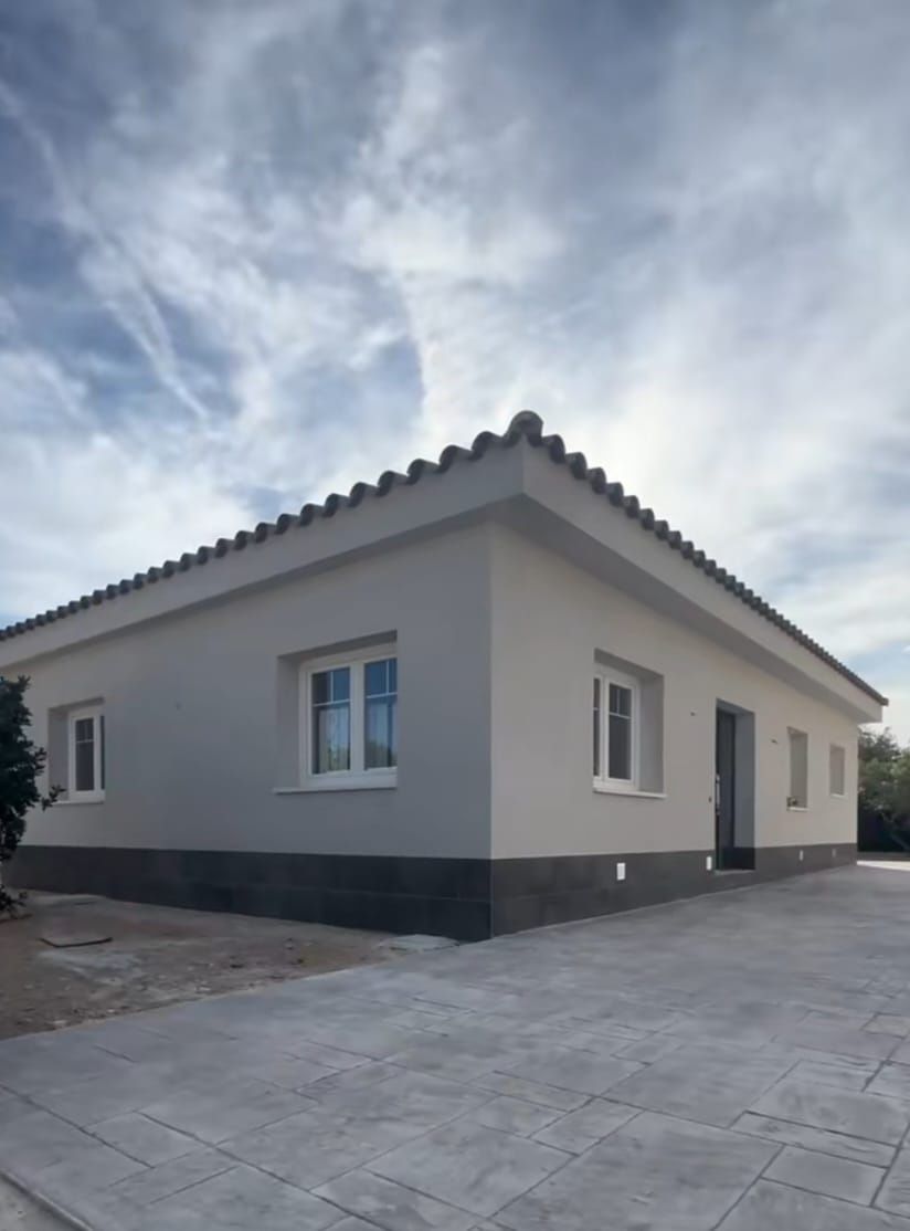 Vista exterior de una casa de una sola planta, de color claro con detalles oscuros y techo de tejas bajo un cielo nublado.