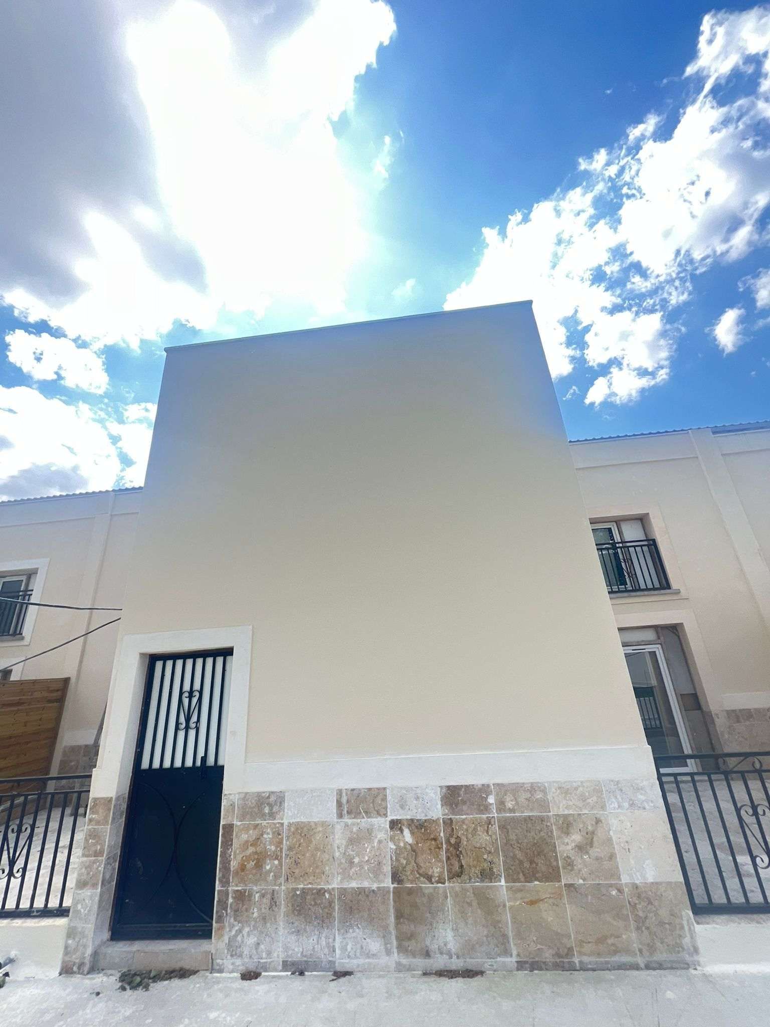 Un edificio de color claro con base de piedra, puerta negra y cielo soleado arriba.