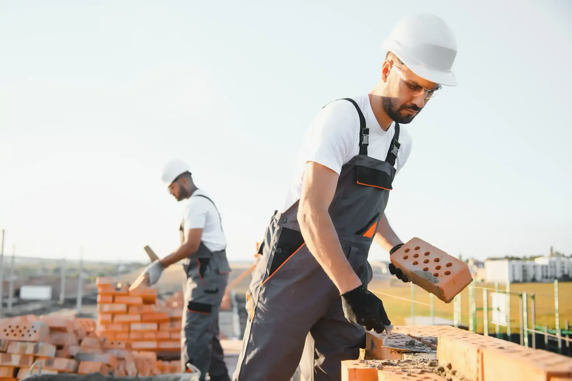Dos obreros de la construcción colocan ladrillos en un tejado; uno de ellos sostiene un ladrillo. Luz solar intensa.