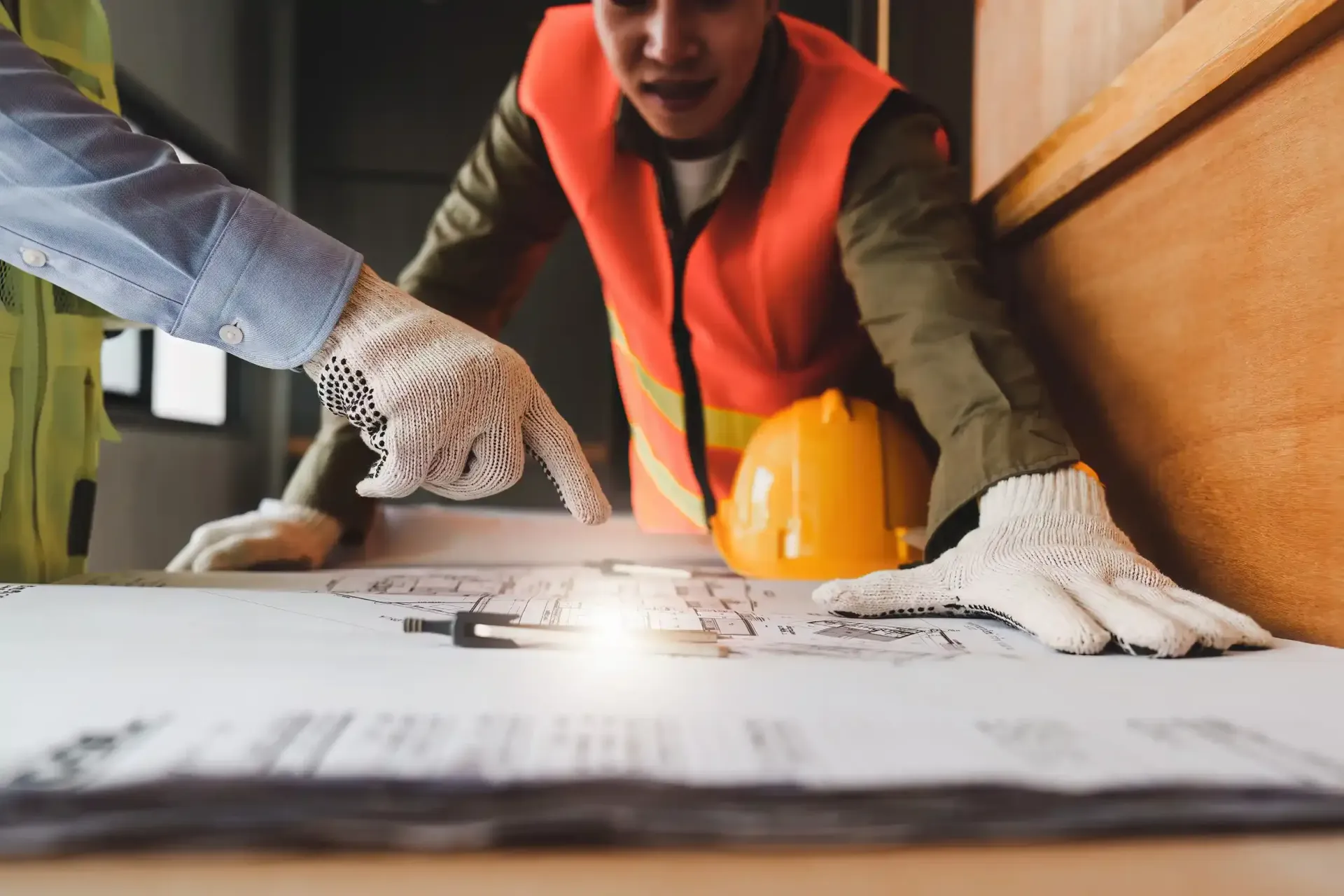 Obreros de la construcción examinan planos sobre una mesa; uno de ellos señala, vistiendo un chaleco naranja.