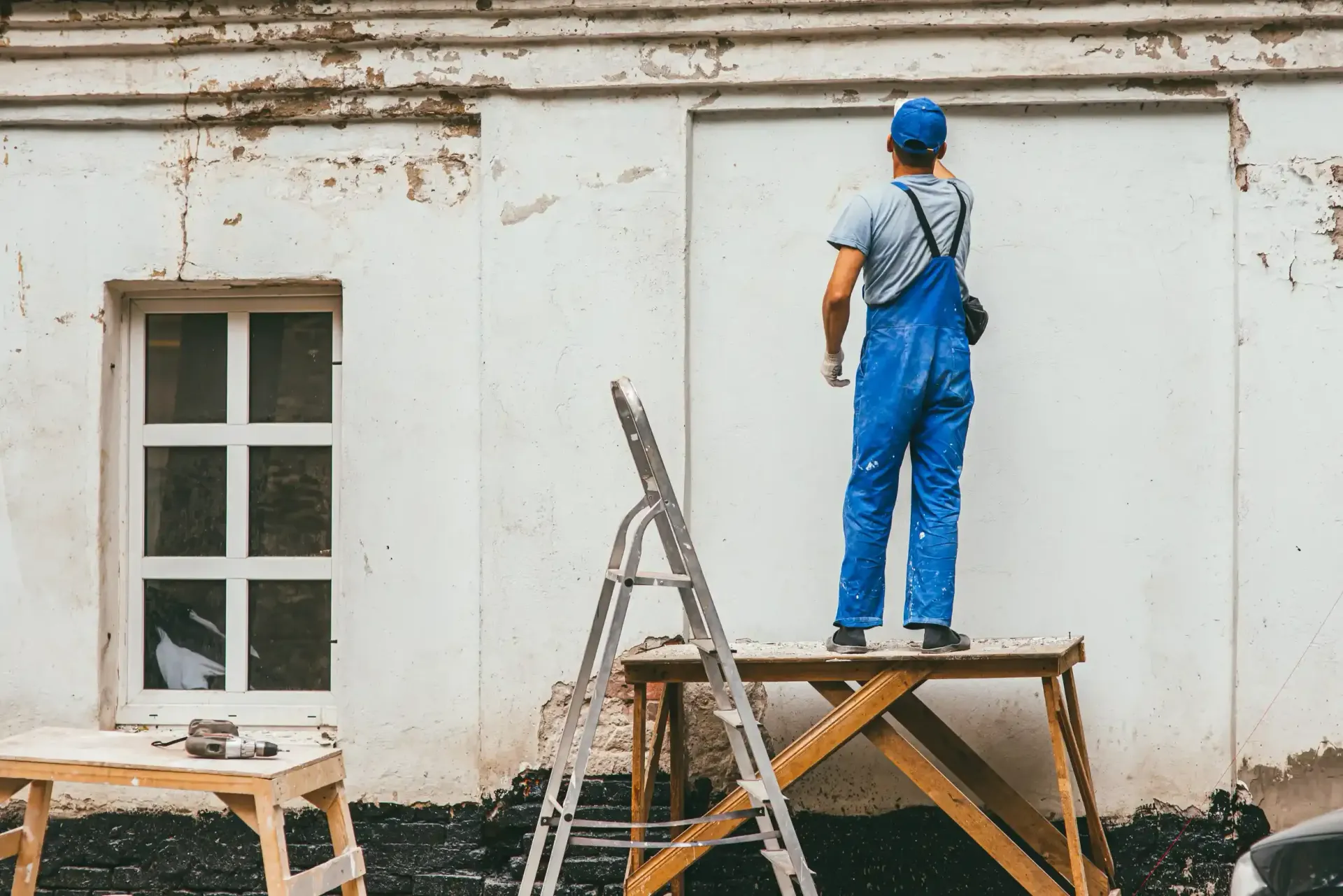 Una persona vestida con un mono azul pinta una pared exterior blanca, de pie sobre una plataforma de madera.