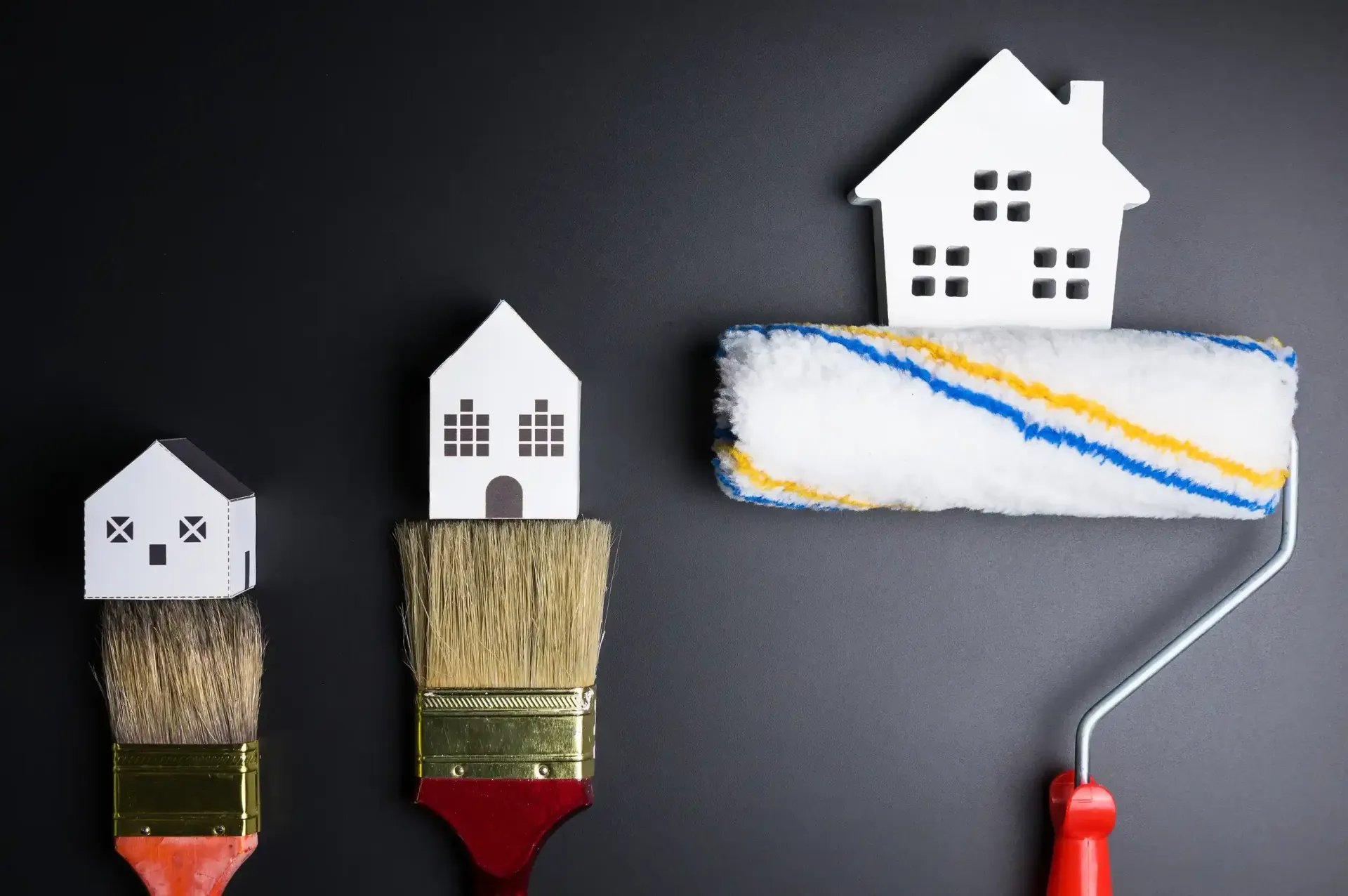 Tres casas de juguete colocadas encima de pinceles y un rodillo de pintura, sobre un fondo negro.