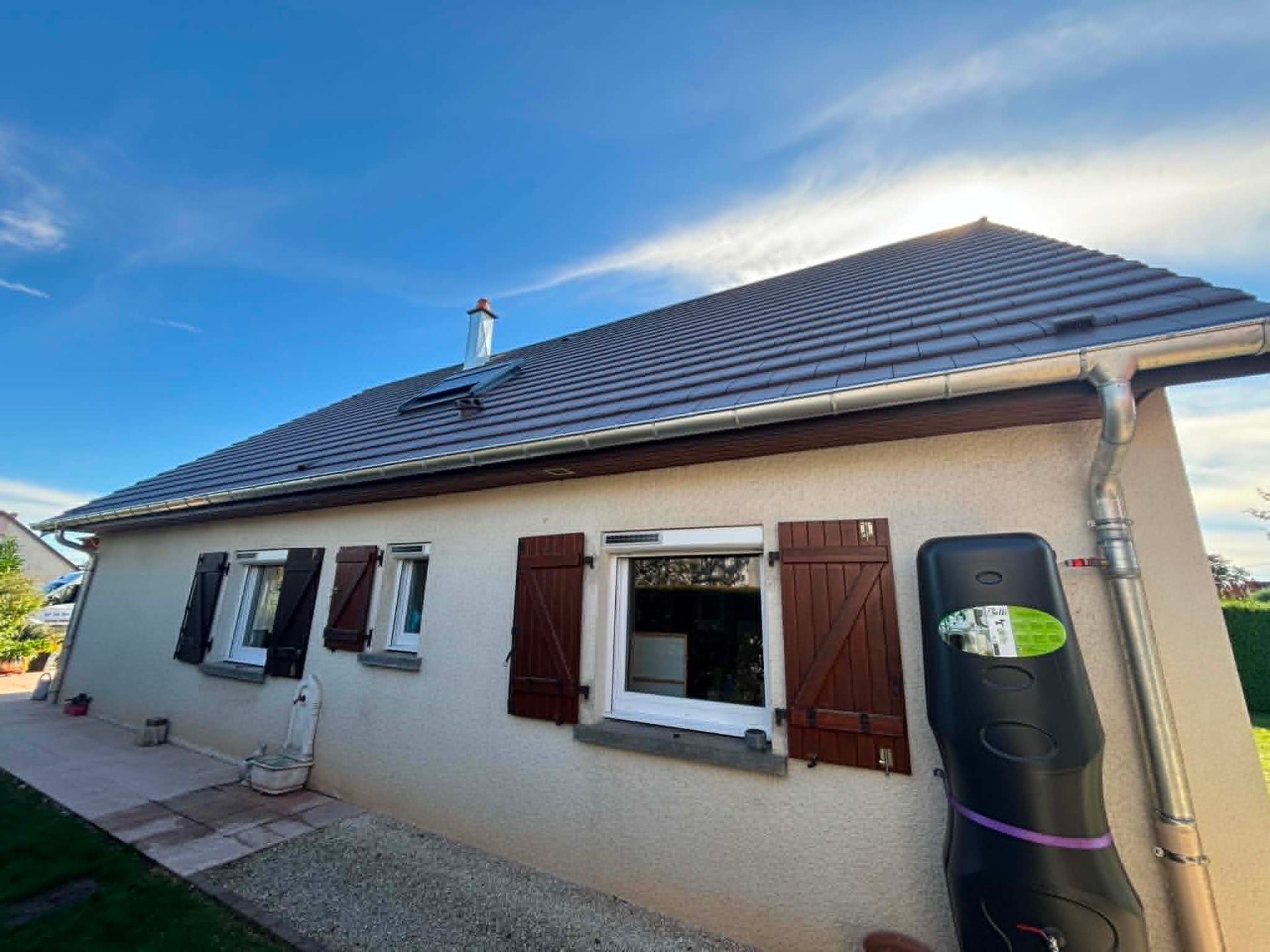 Maison beige de plain-pied avec des volets marron et un réservoir d'eau noir sous un ciel bleu