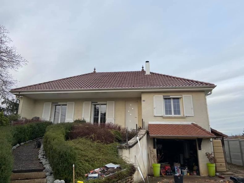 Maison beige à deux étages avec toit de tuiles rouges, marches d'entrée et garage ouvert au milieu de buissons envahissants.