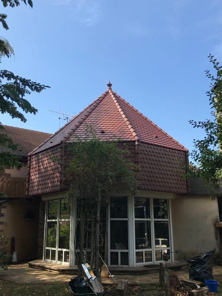 Petite maison au toit de tuiles rouges et aux grandes fenêtres, partiellement recouverte de vigne, dans une cour arborée.