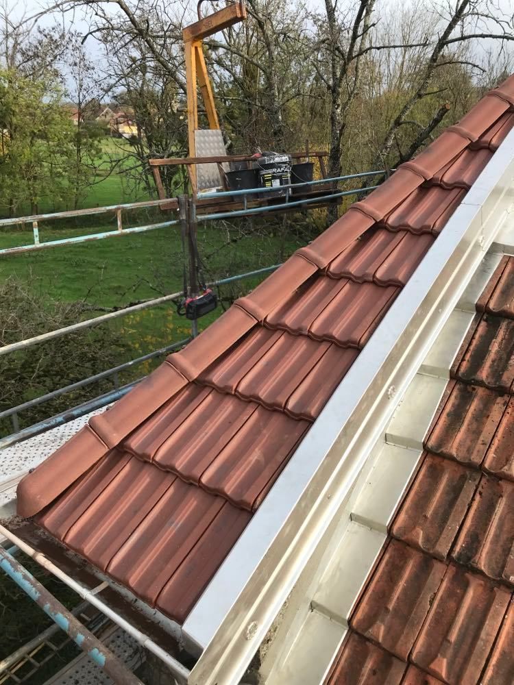 Un couvreur pose des tuiles rouges sur un toit en pente, à côté d'une gouttière blanche, avec un échafaudage et des arbres en arrière-plan.