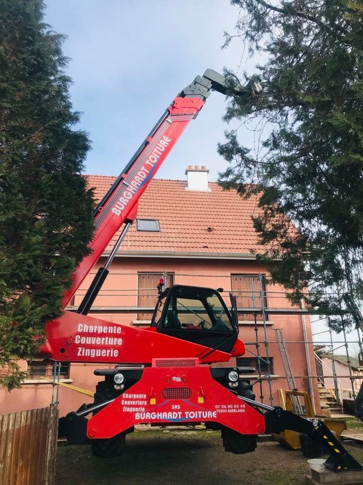 Une nacelle élévatrice rouge de chantier se trouve à côté d'une maison, avec une flèche déployée entre les arbres et un toit de tuiles.