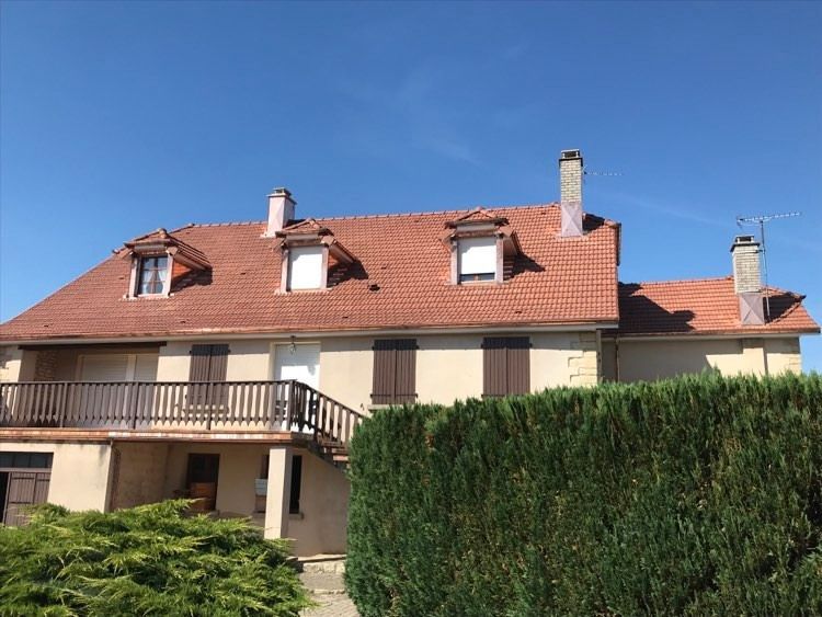Maison à deux étages avec un toit de tuiles rouges, une façade beige, un balcon et une haute haie sous un ciel bleu clair.