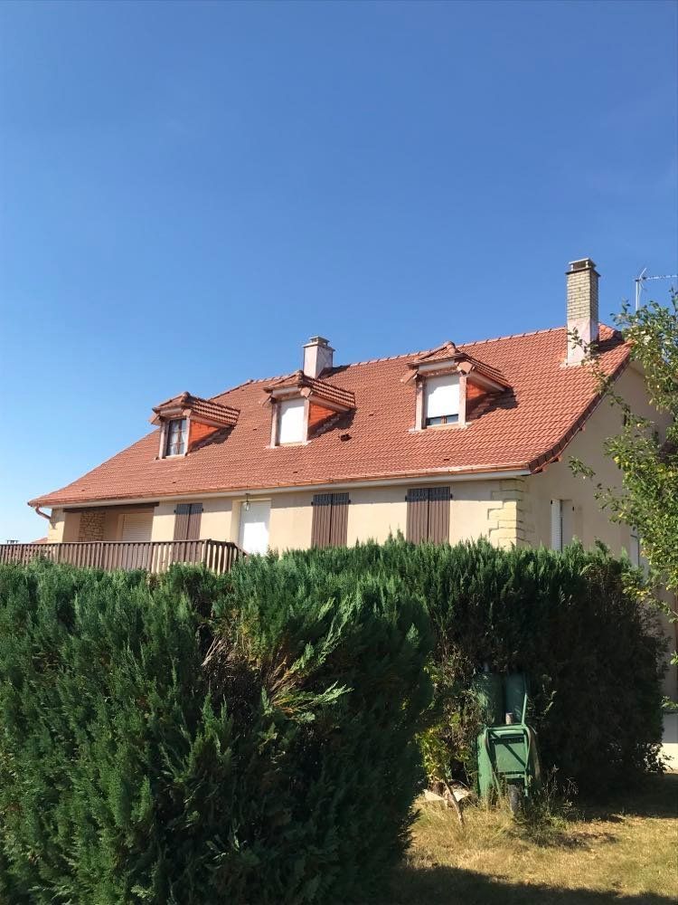 Maison au toit rouge avec lucarnes, dissimulée derrière de hautes haies vertes sous un ciel d'un bleu limpide.