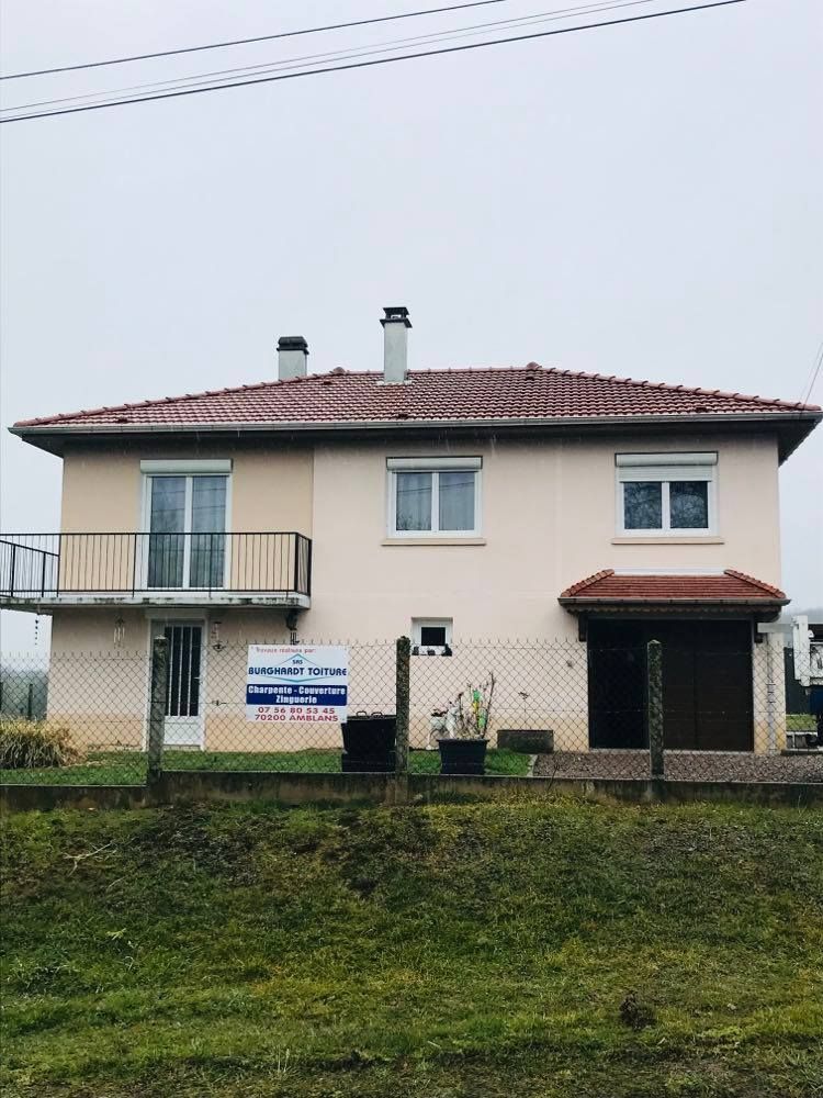 Maison à deux étages avec toit rouge et balcon, vue depuis une cour herbeuse par temps nuageux