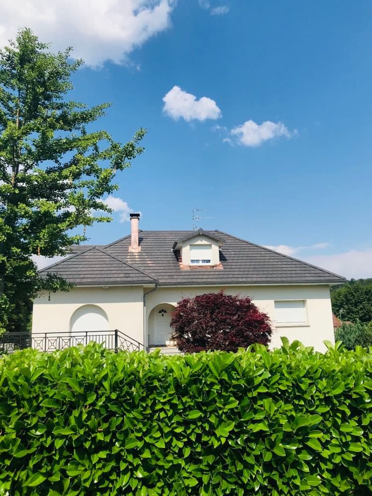 Maison au toit gris et lucarne dissimulée derrière une haie verte sous un ciel d'un bleu éclatant