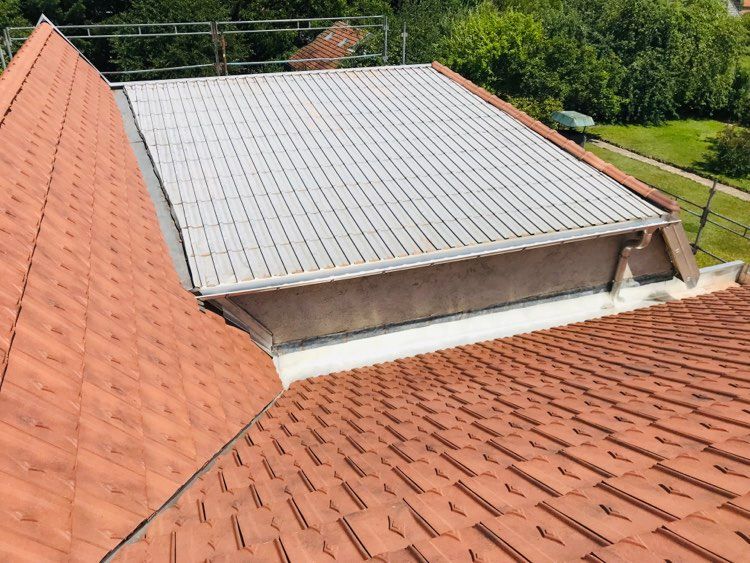 Toiture recouverte de tuiles rouges entourant une section de toit en tôle ondulée grise et entourée de verdure.