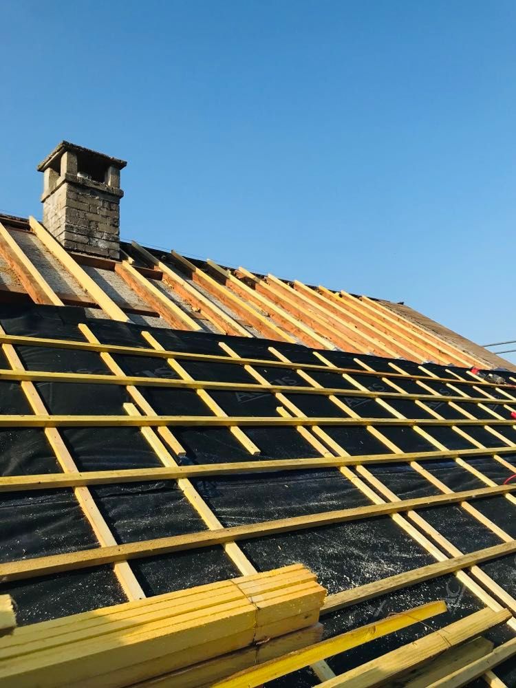 Toiture en construction avec des liteaux en bois jaunes sur une sous-couche noire et une petite cheminée se détachant sur un ciel bleu