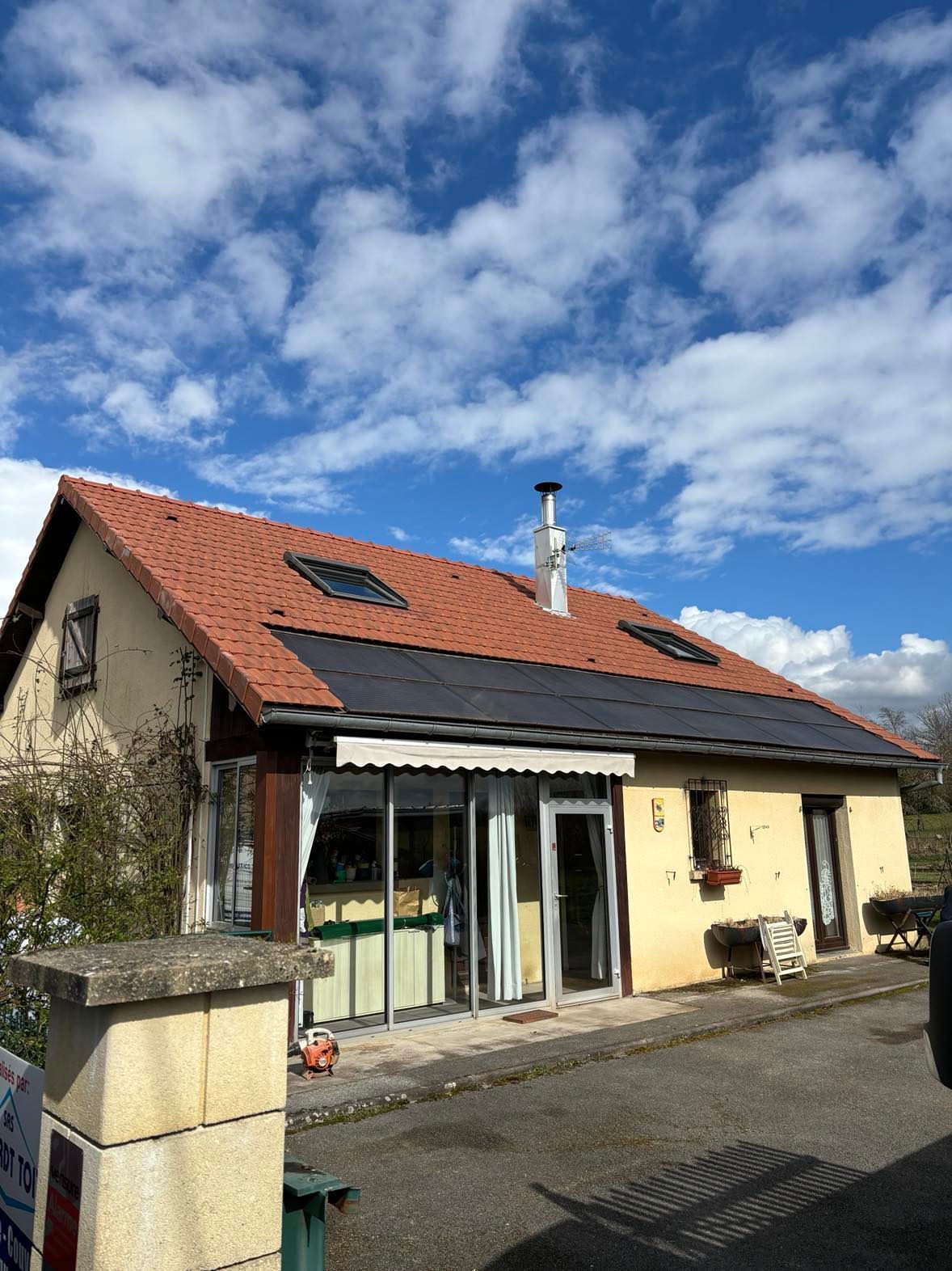 Petite maison au toit de tuiles rouges, avec puits de lumière et patio sous un ciel bleu nuageux.