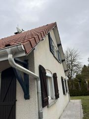 Vue latérale d'une maison couleur crème avec un toit de tuiles rouges, des volets sombres et une allée pavée sous un ciel nuageux