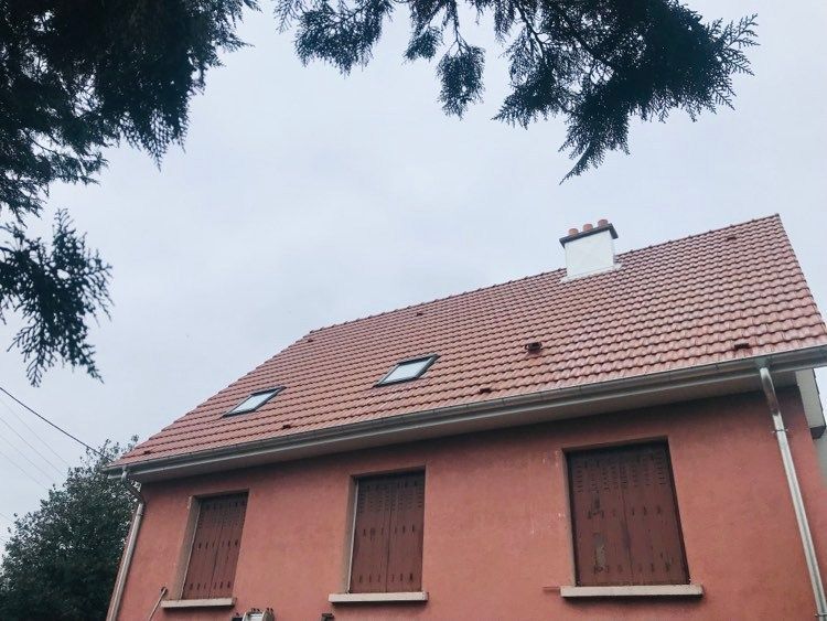 Maison rose au toit de tuiles rouges et aux fenêtres à volets, vue d'en bas à travers les branches des arbres