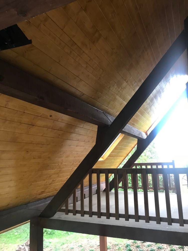 Plafond en bois incliné et poutres sombres au-dessus d'une balustrade de balcon, avec une vive lumière du jour à l'extérieur.