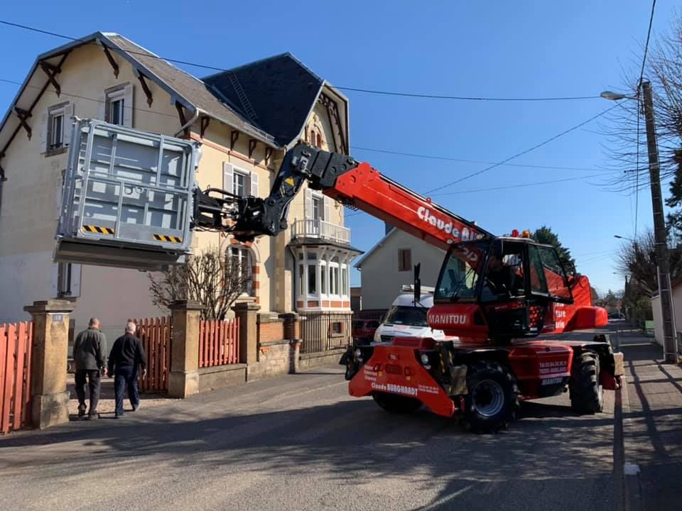 Camion-grue rouge soulevant un balcon dans une rue adjacente à un immeuble résidentiel.