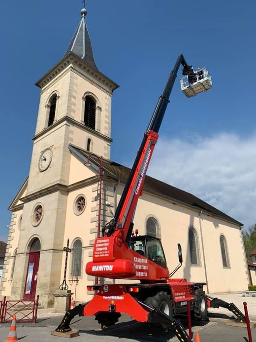 Une nacelle élévatrice rouge soulève un ouvrier à côté d'une église beige avec un haut clocher sous un ciel bleu.