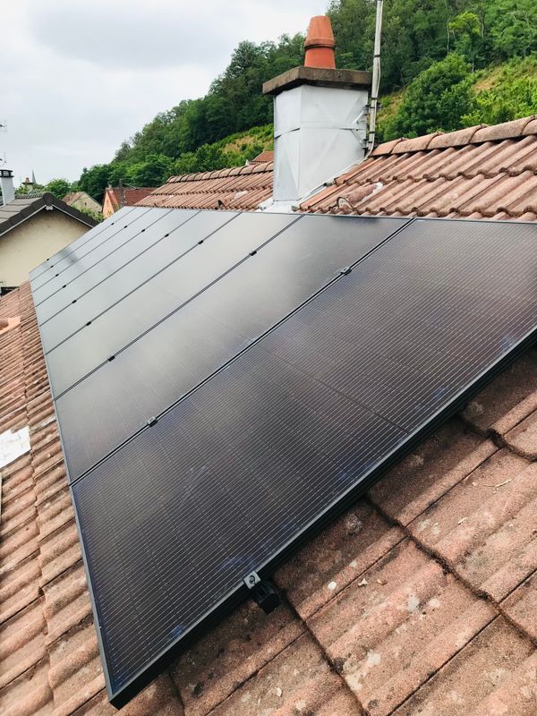Des panneaux solaires installés sur un toit de tuiles rouges à côté d'une cheminée, avec des collines verdoyantes en arrière-plan.