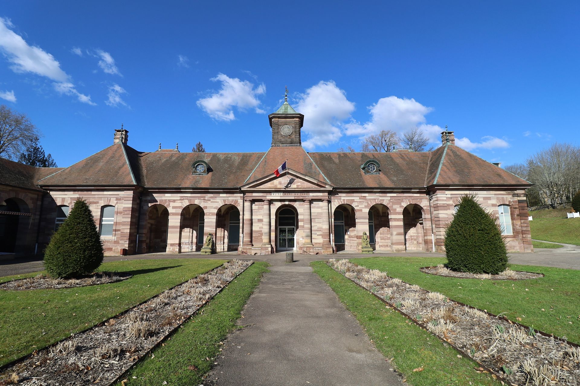 Bâtiment en pierre avec entrée centrale et allée paysagée sous un ciel bleu