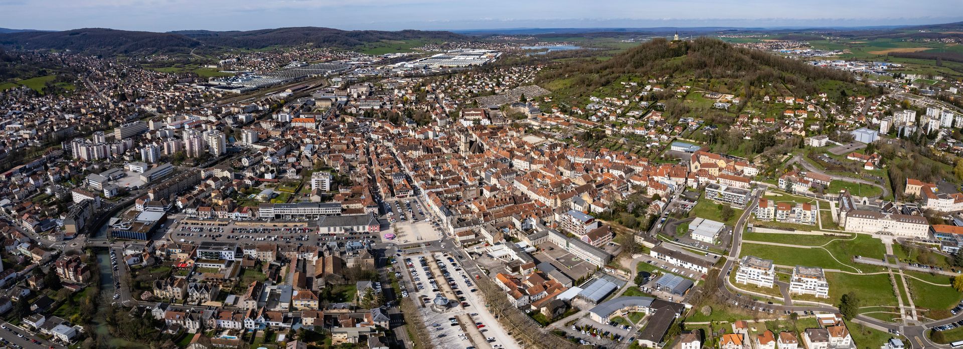 Vue aérienne d'une ville dense avec des immeubles aux toits rouges, des rues et des collines verdoyantes à l'horizon.