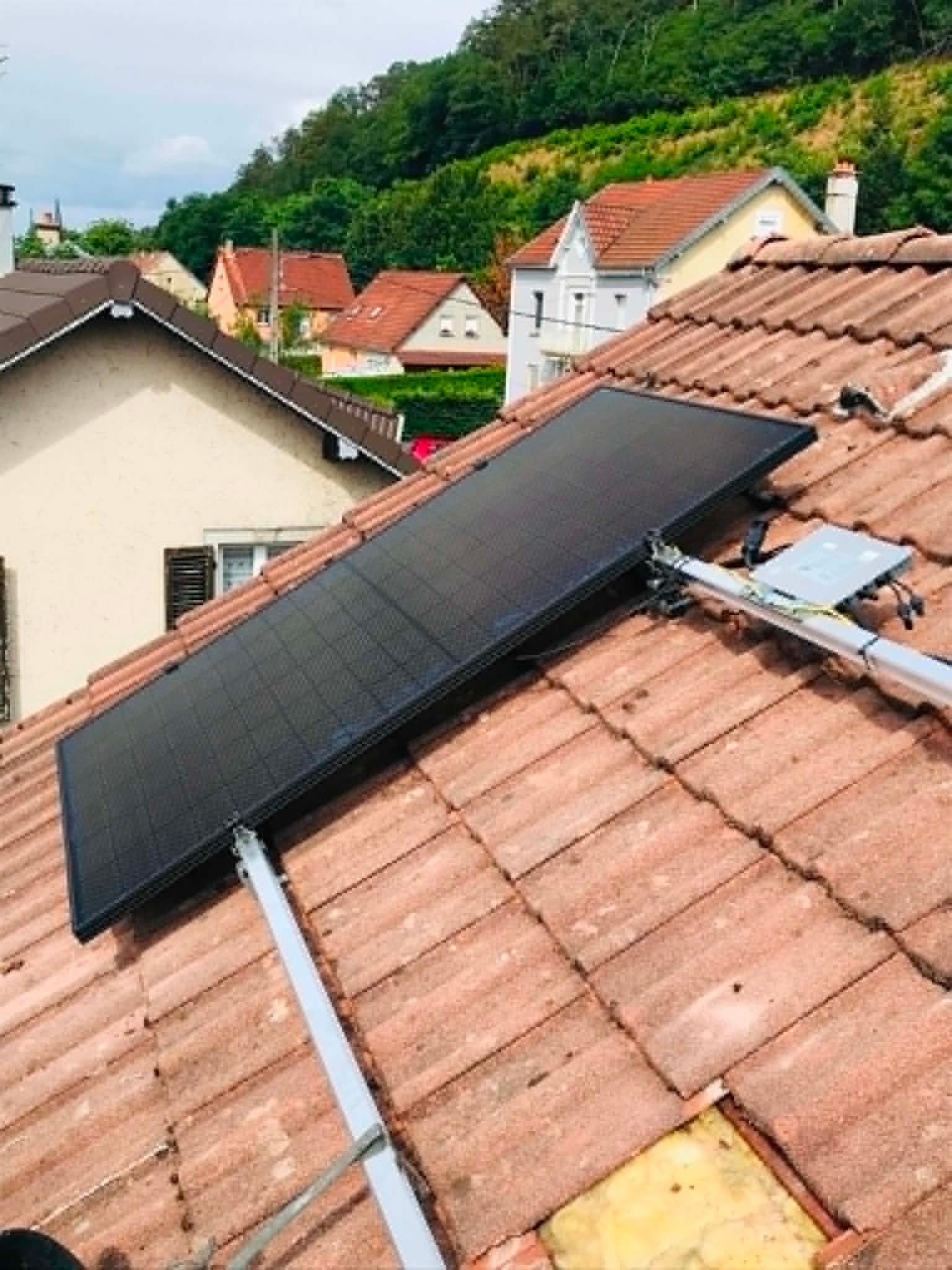 Panneau solaire noir installé sur un toit de tuiles rouges, avec des maisons et une colline verdoyante en arrière-plan.